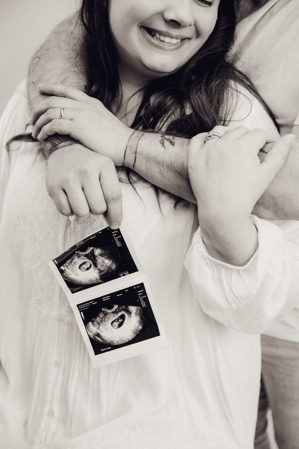 Black and white close-up of a smiling pregnant woman holding ultrasound images while being embraced by partner.