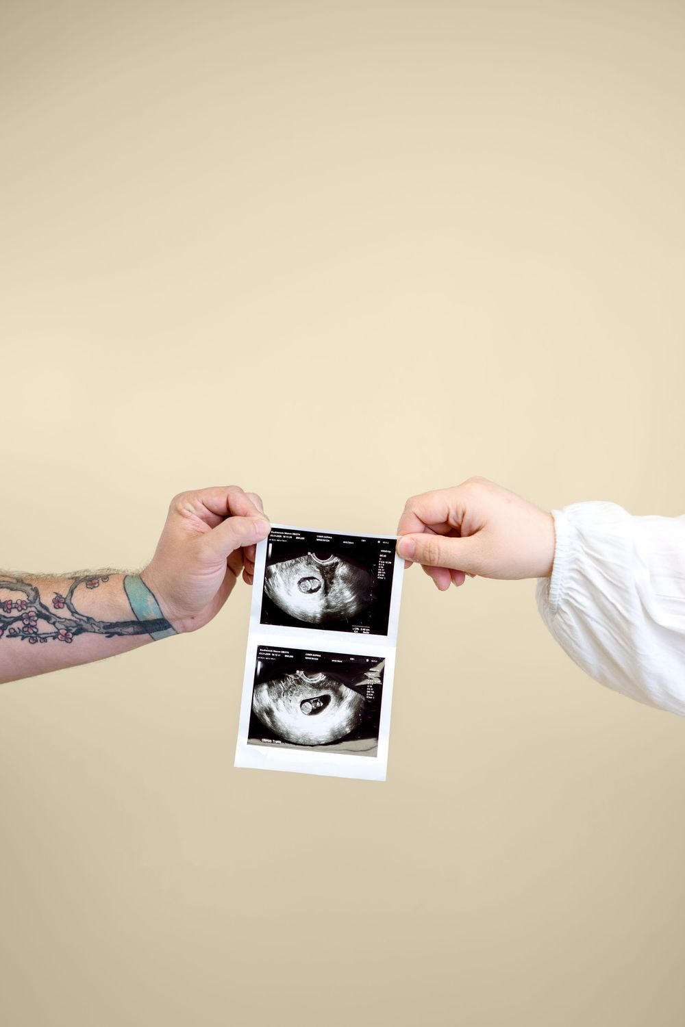Tattooed and plain hands holding baby ultrasound scans on beige background.