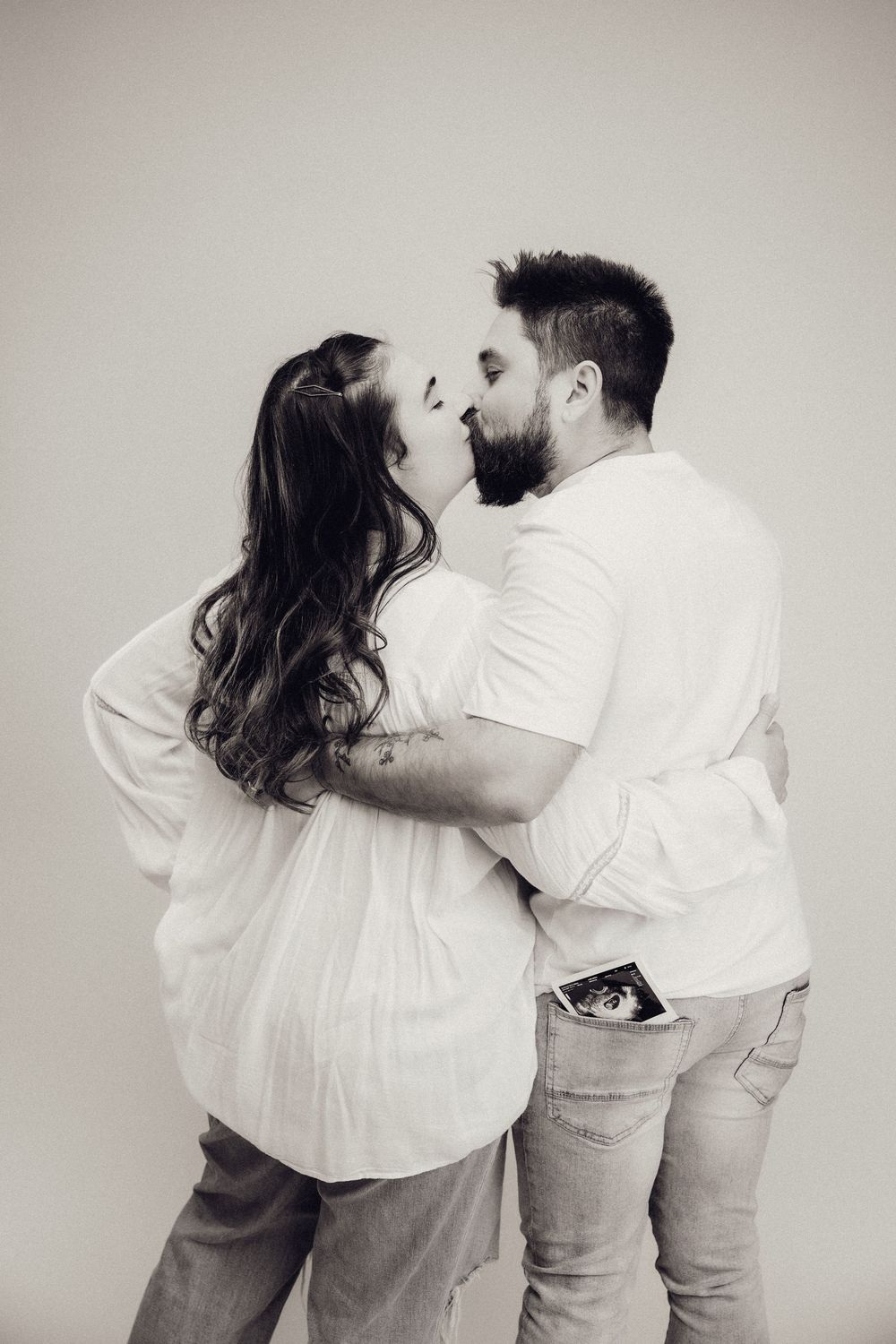 Black and white photo of a couple kissing while man holds ultrasound scan in his back pocket.