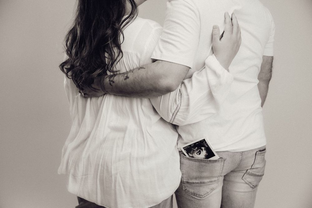 Black and white rear view of a pregnant couple standing together with ultrasound photo in mans back pocket.