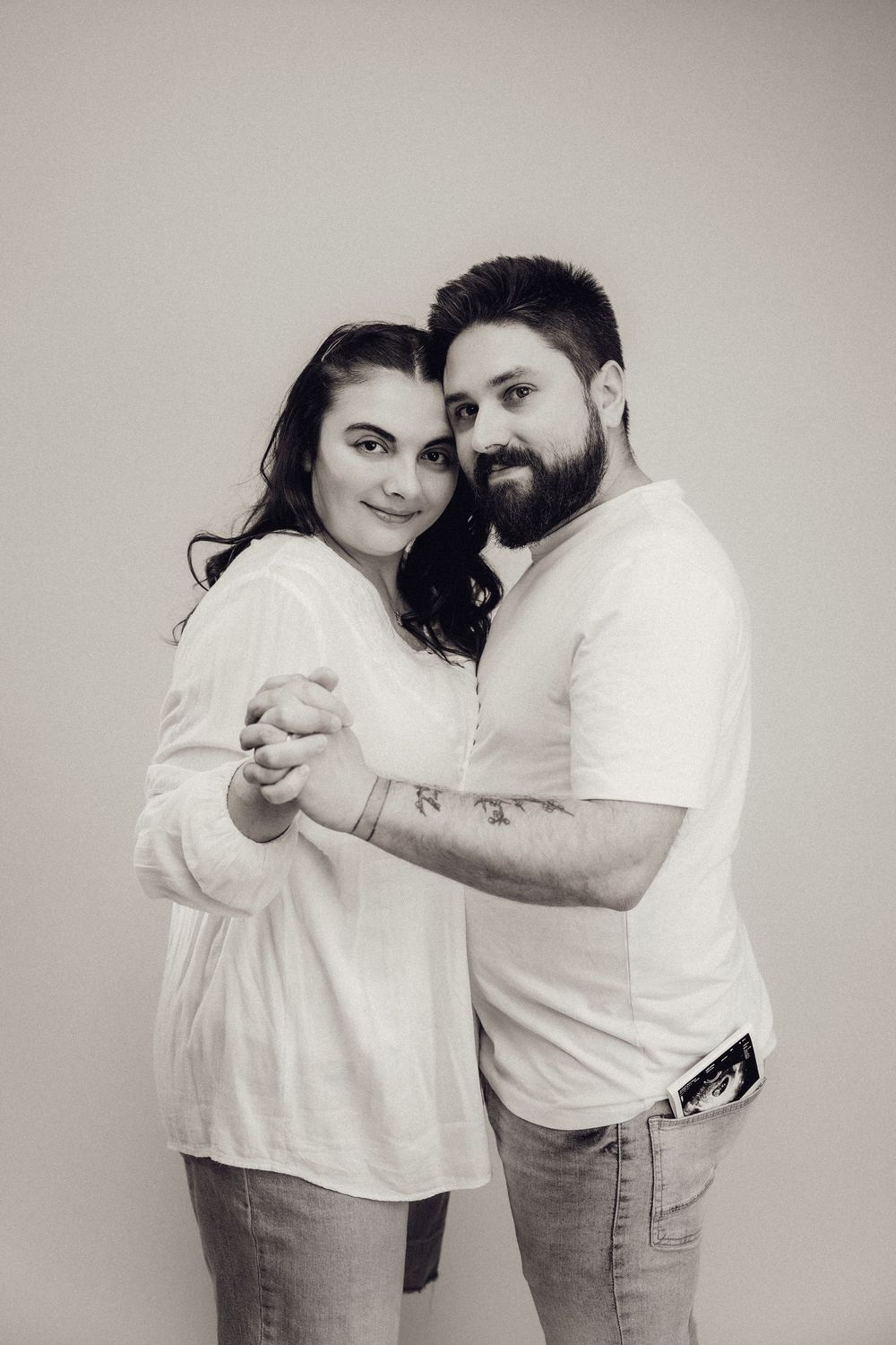 Black and white portrait of a smiling couple holding hands together, man with ultrasound scan in pocket.