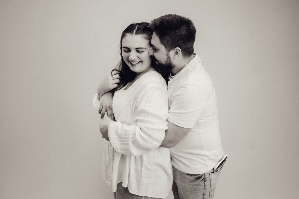 Black and white photo of a laughing couple embracing closely during a maternity photo session.