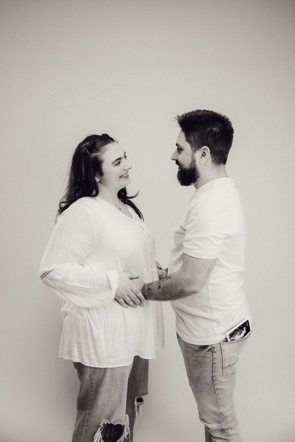 Black and white photo of a smiling pregnant couple in white tops and jeans holding her baby bump together.