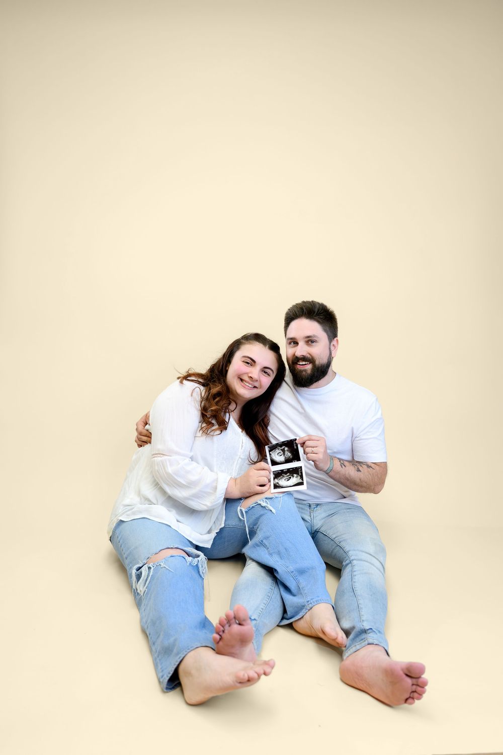 Happy couple sitting on floor holding ultrasound photos, smiling at camera.