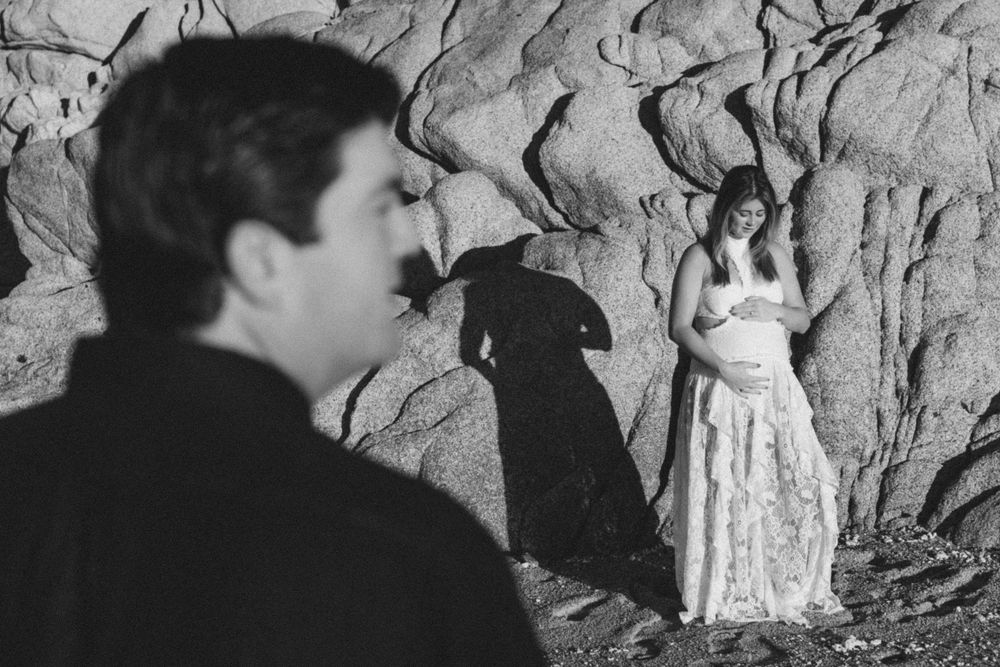 Black and white photo of a couple near rocky cliffs, woman in flowing dress stands illuminated while man watches from foreground.