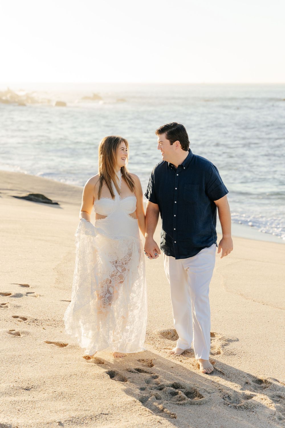 Couple walking hand-in-hand on a sandy beach at sunset, woman in white dress, man in navy shirt and white pants.