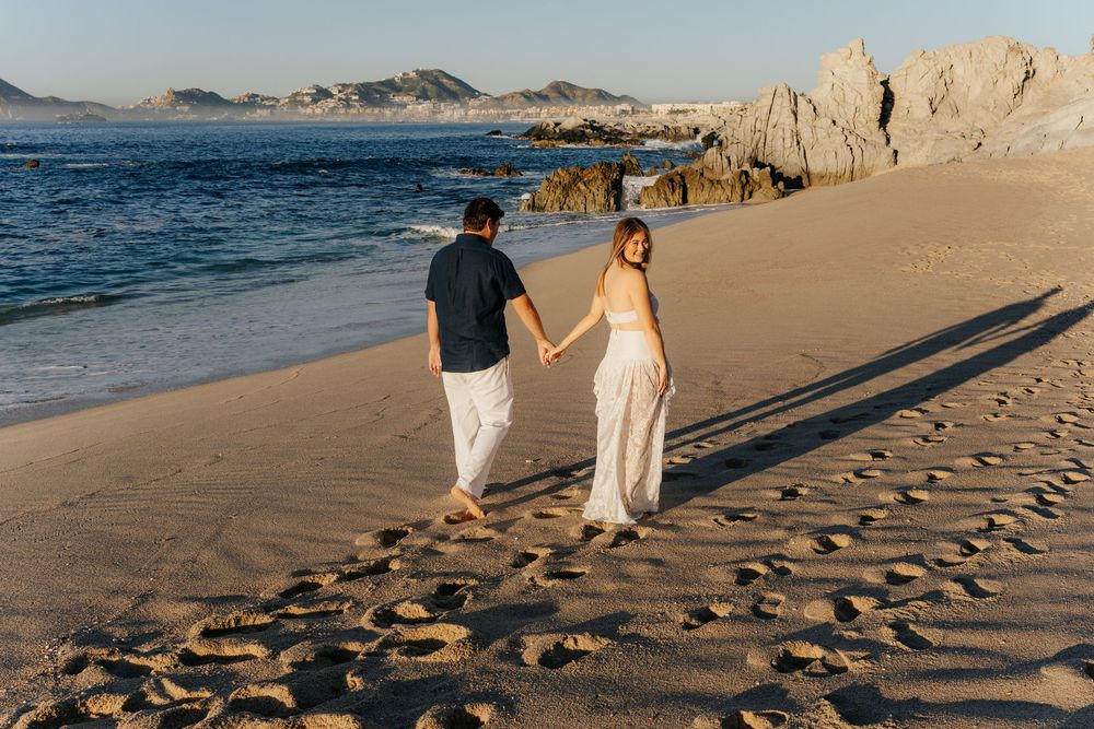 Couple holding hands walking on sandy beach at sunset with rocky cliffs and ocean waves in background.