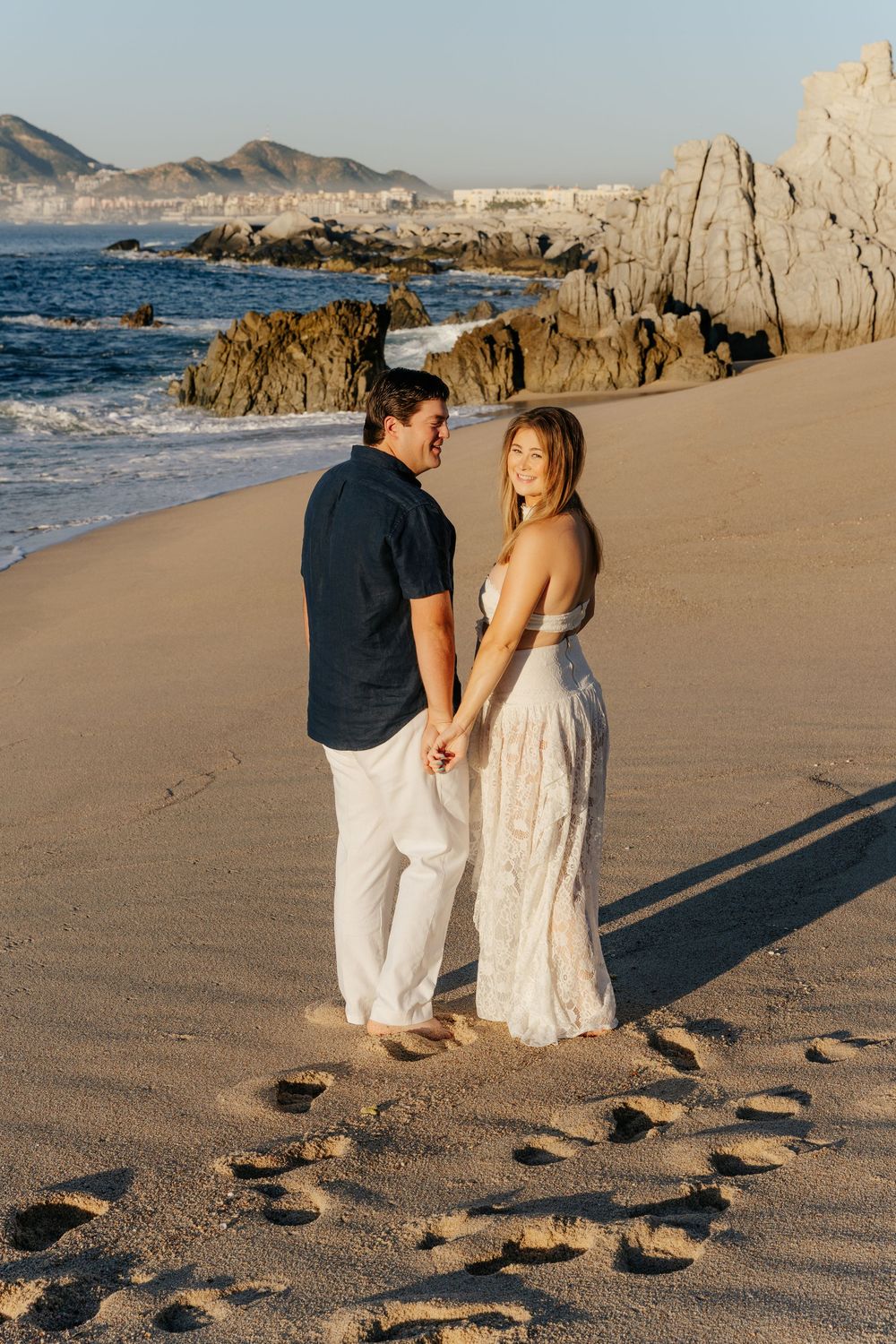 Couple holding hands on a sandy beach at golden hour, wearing white and navy, with rocky coastline in the background.