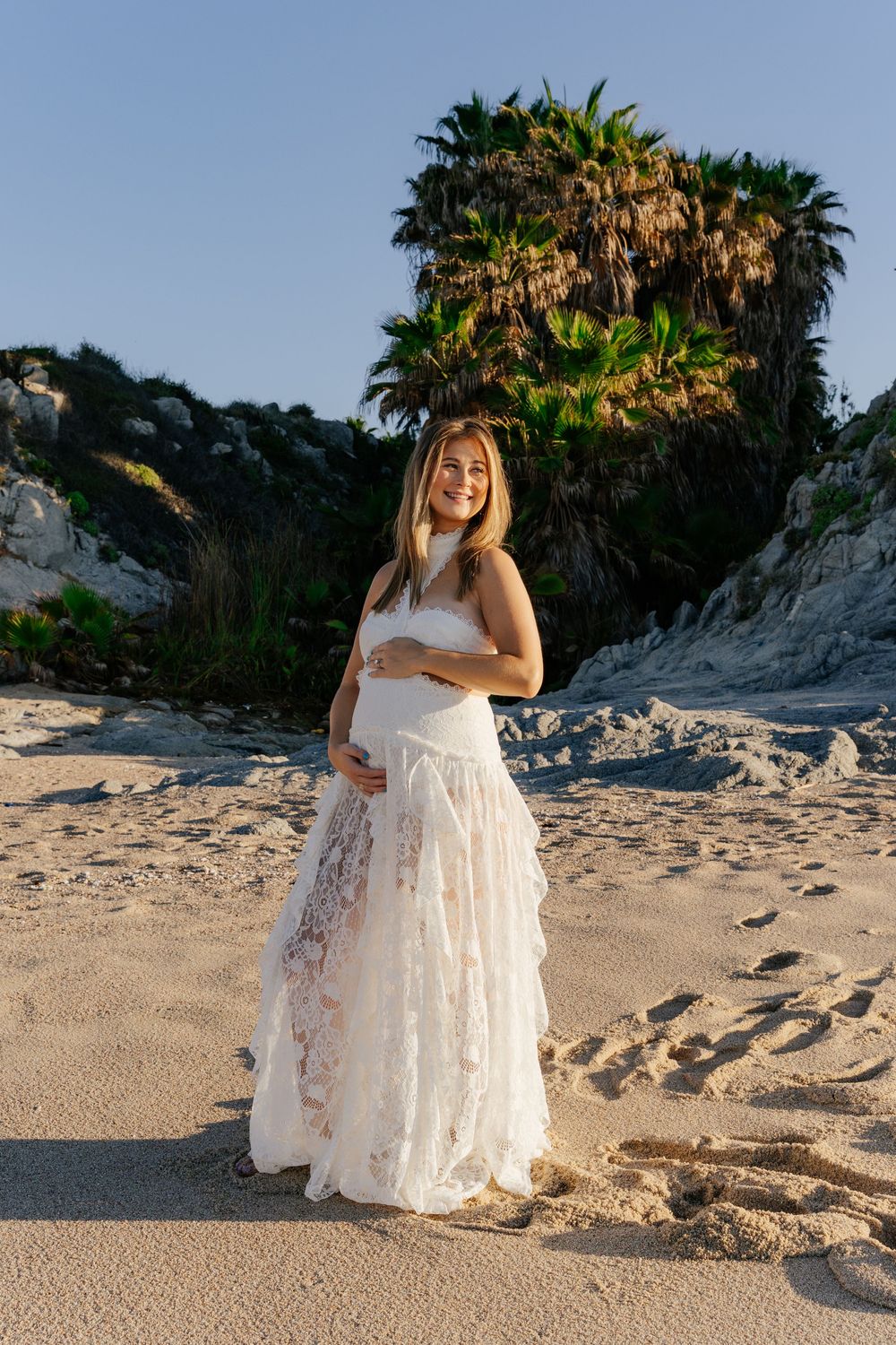 Pregnant woman in white lace dress smiling on sandy beach with palm trees and cliffs in golden hour light.
