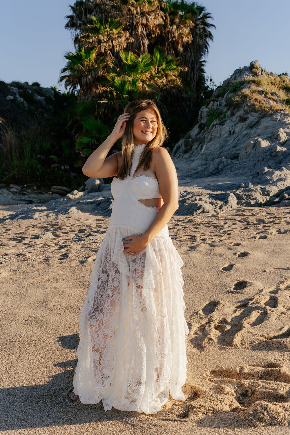 Woman in white lace dress smiling on sandy beach with palm trees and rocky cliffs in golden hour light.