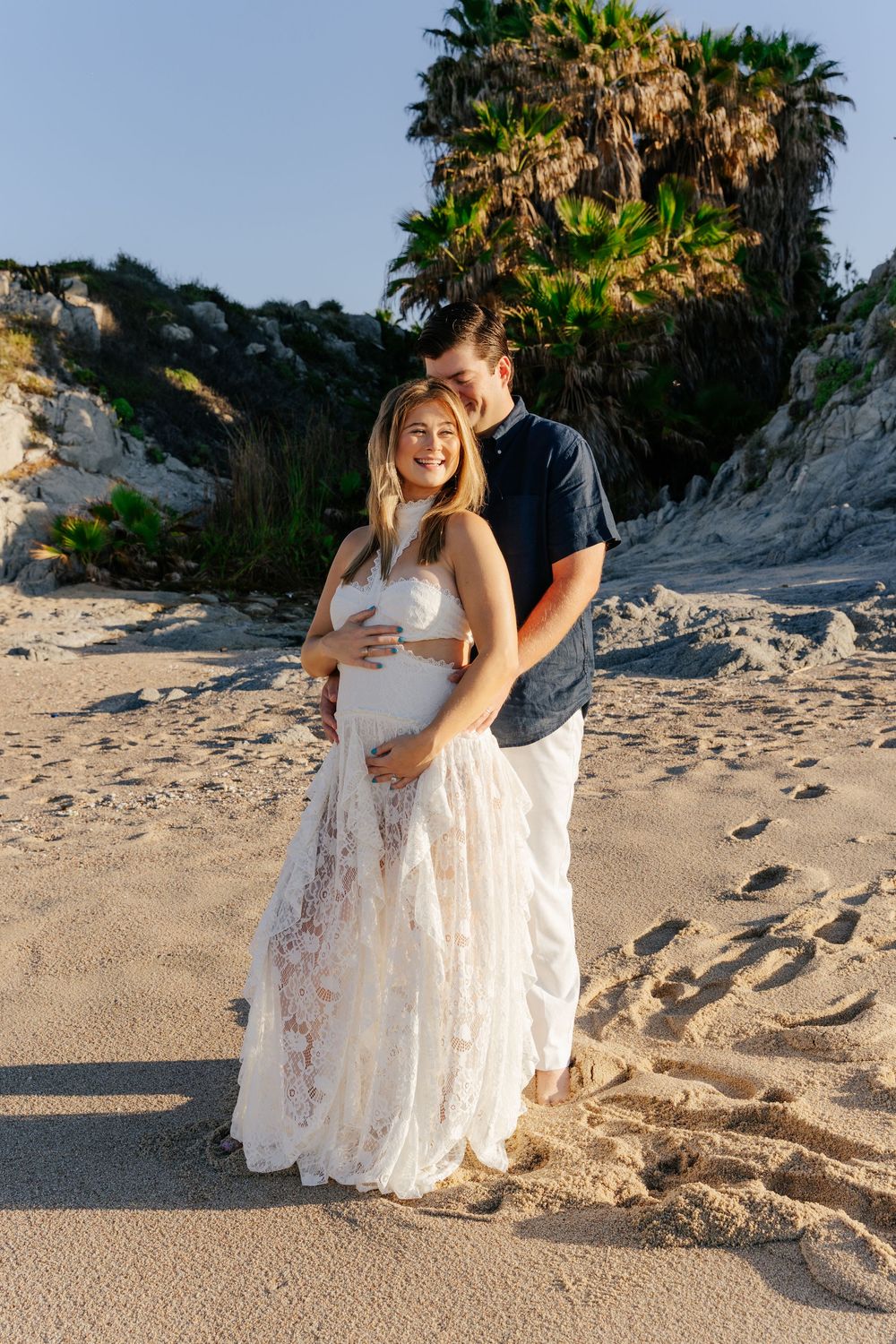 Couple poses on sandy beach at sunset, pregnant woman in white lace gown, man in navy shirt, palm trees behind them.