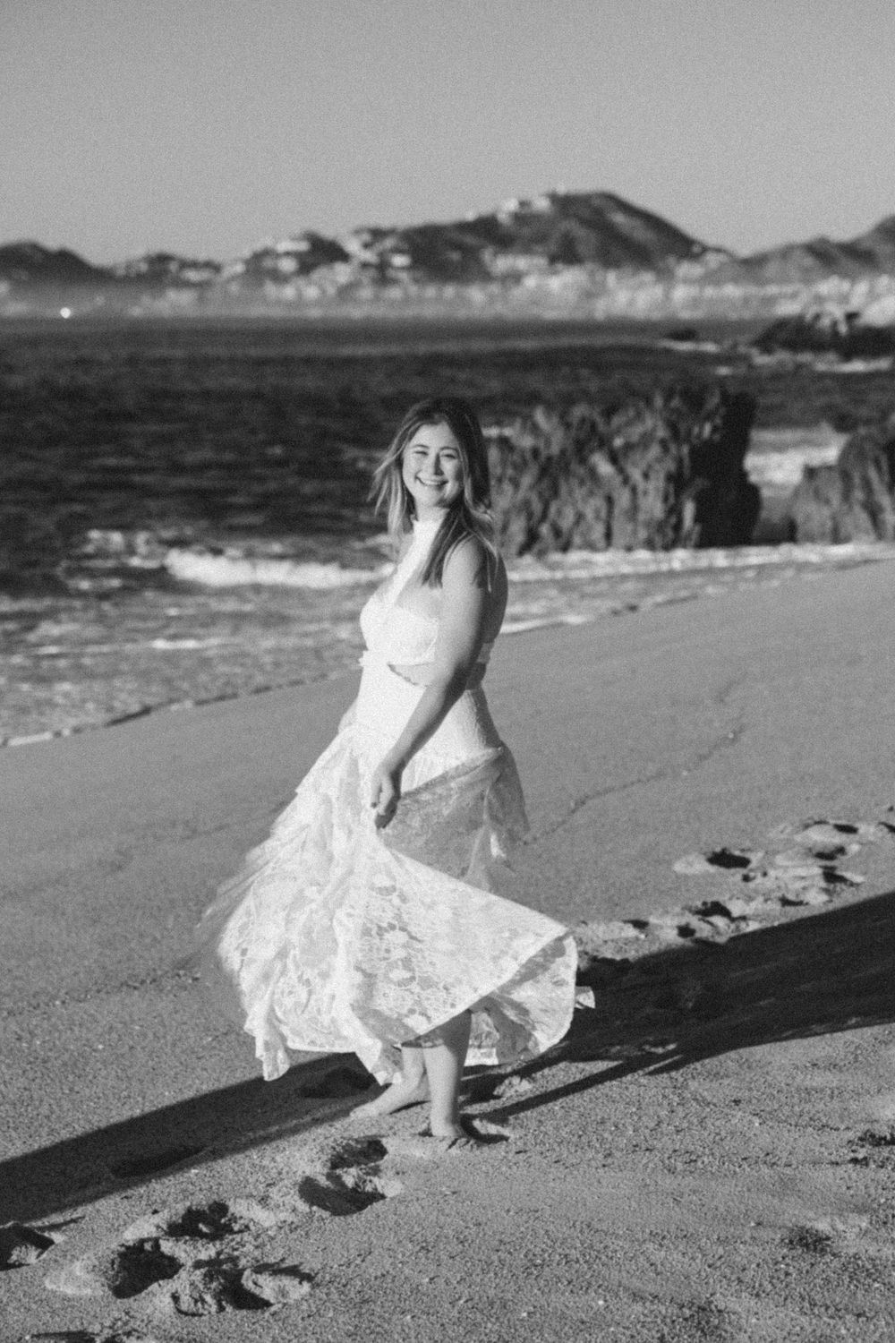 Black and white photo of a young woman in a lace dress walking barefoot on a sandy beach with ocean waves behind her.