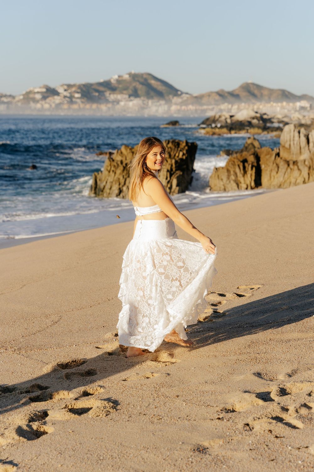 Woman in white lace dress walking on sandy beach with rocky coastline and ocean waves in background.