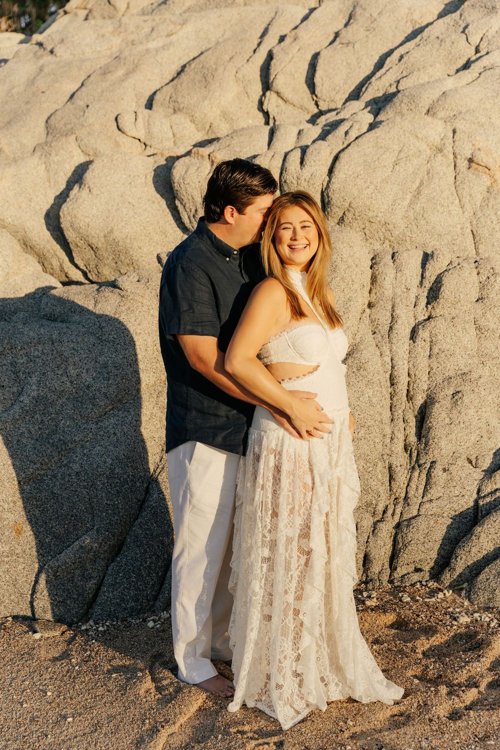Couple poses against large rocks at golden hour, woman in white lace maternity gown cradling baby bump at beach.