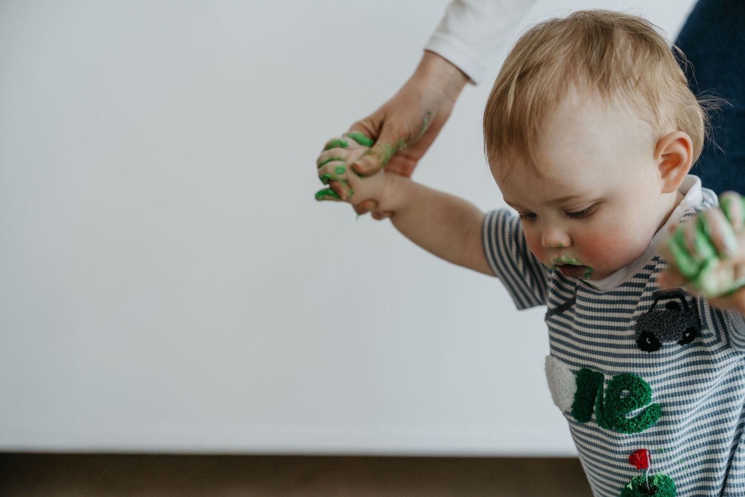 Toddler with green paint on hands being helped to stand by adult, wearing striped shirt.