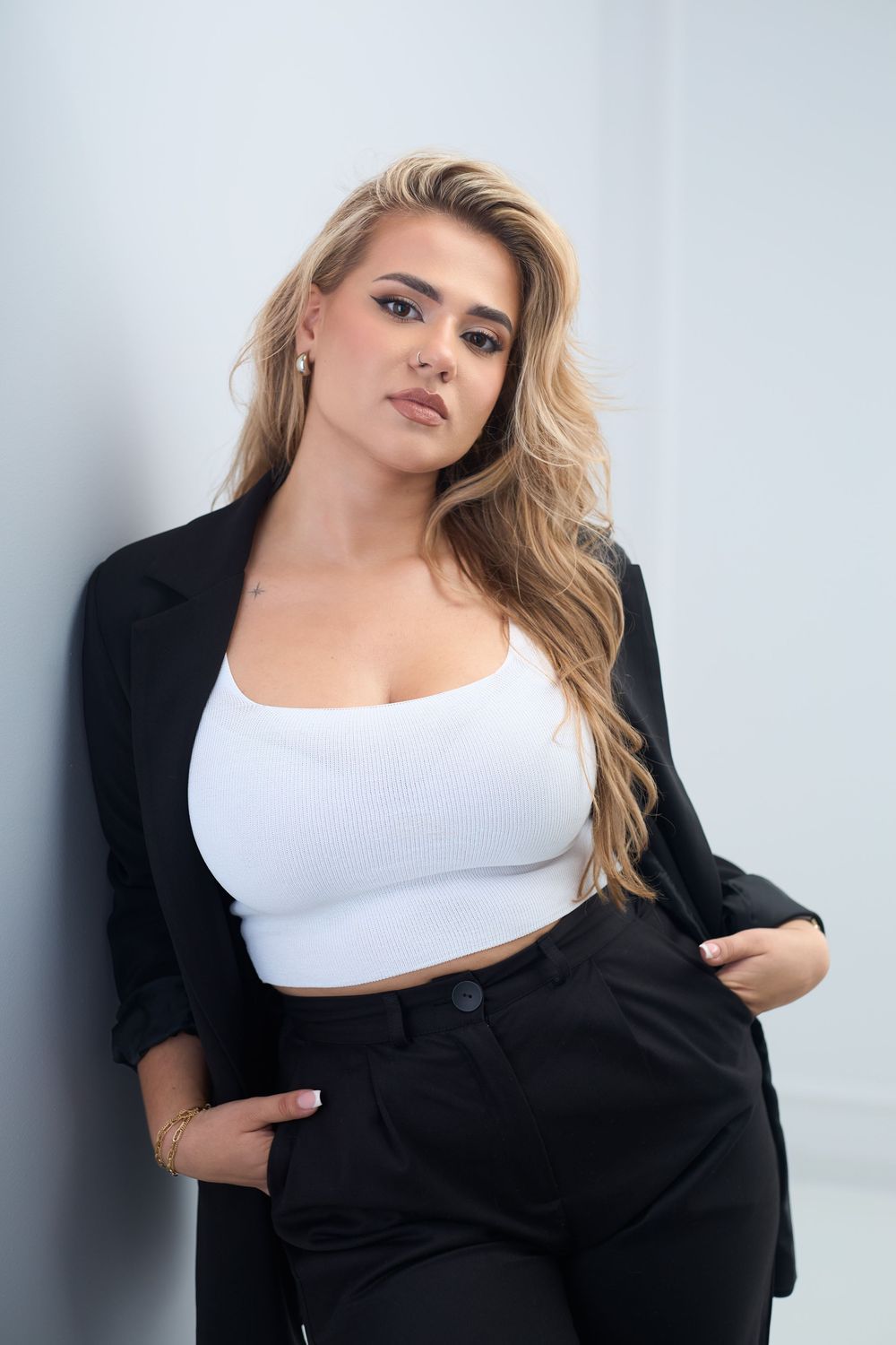 Blonde woman in white crop top, black blazer and pants posing confidently against a light gray wall.