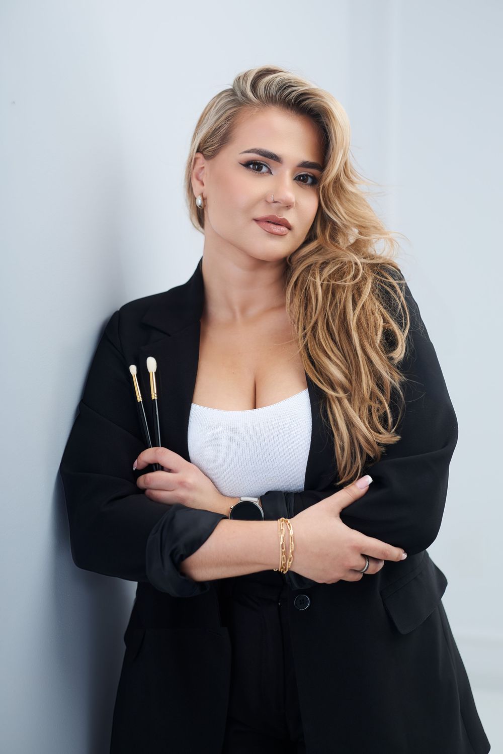 Blonde woman in black blazer holding makeup brushes, wearing gold jewelry, posing against white wall.