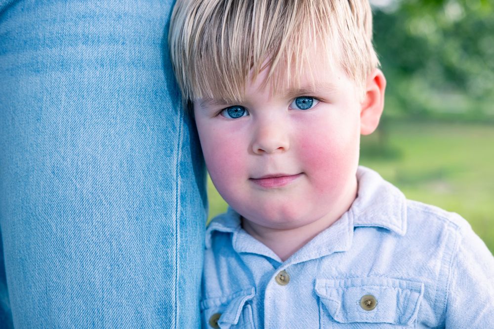 Adorable blonde toddler boy with blue eyes leaning against an adult's leg outdoors.