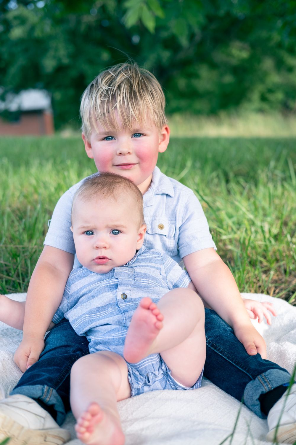 Two young brothers sitting on a blanket outdoors, older blonde boy holding baby sibling, green grass background.