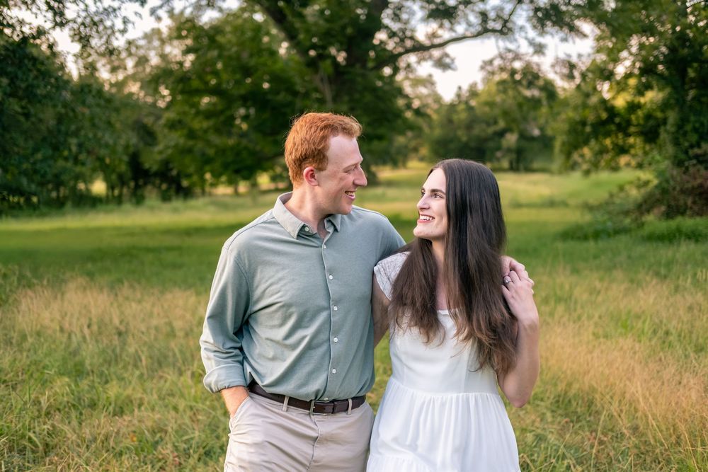 A smiling couple walks through a lush green field, the man in a sage shirt and woman in a white dress.