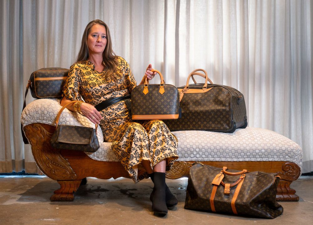 Woman in a gold patterned dress sits on an ornate cushioned bench surrounded by several vintage monogram leather handbags and travel bags.