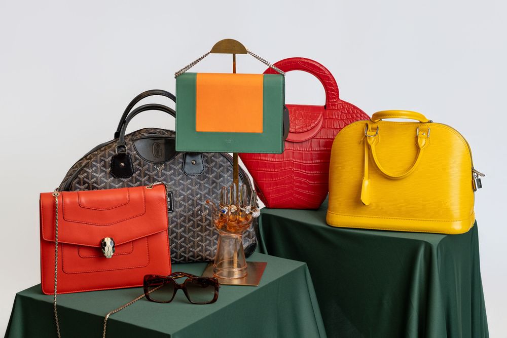 Collection of luxury designer handbags in red, yellow, orange, and gray displayed on green draped pedestals with sunglasses.