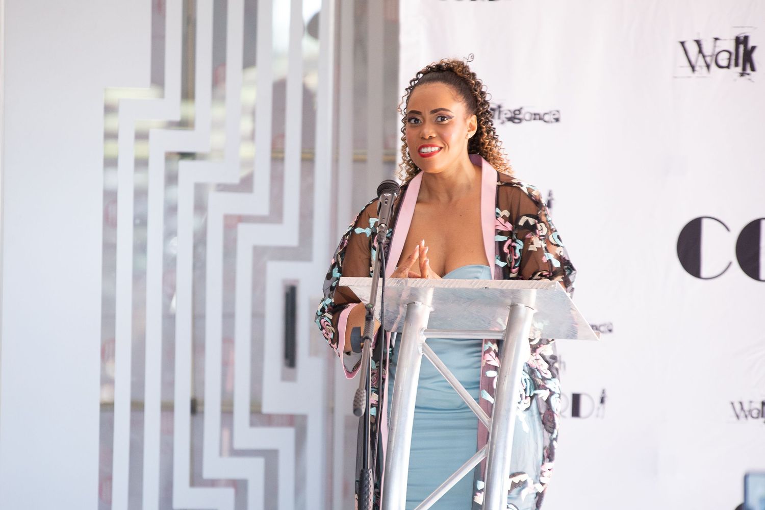 Woman in floral top speaking at podium during outdoor event with decorative geometric backdrop.