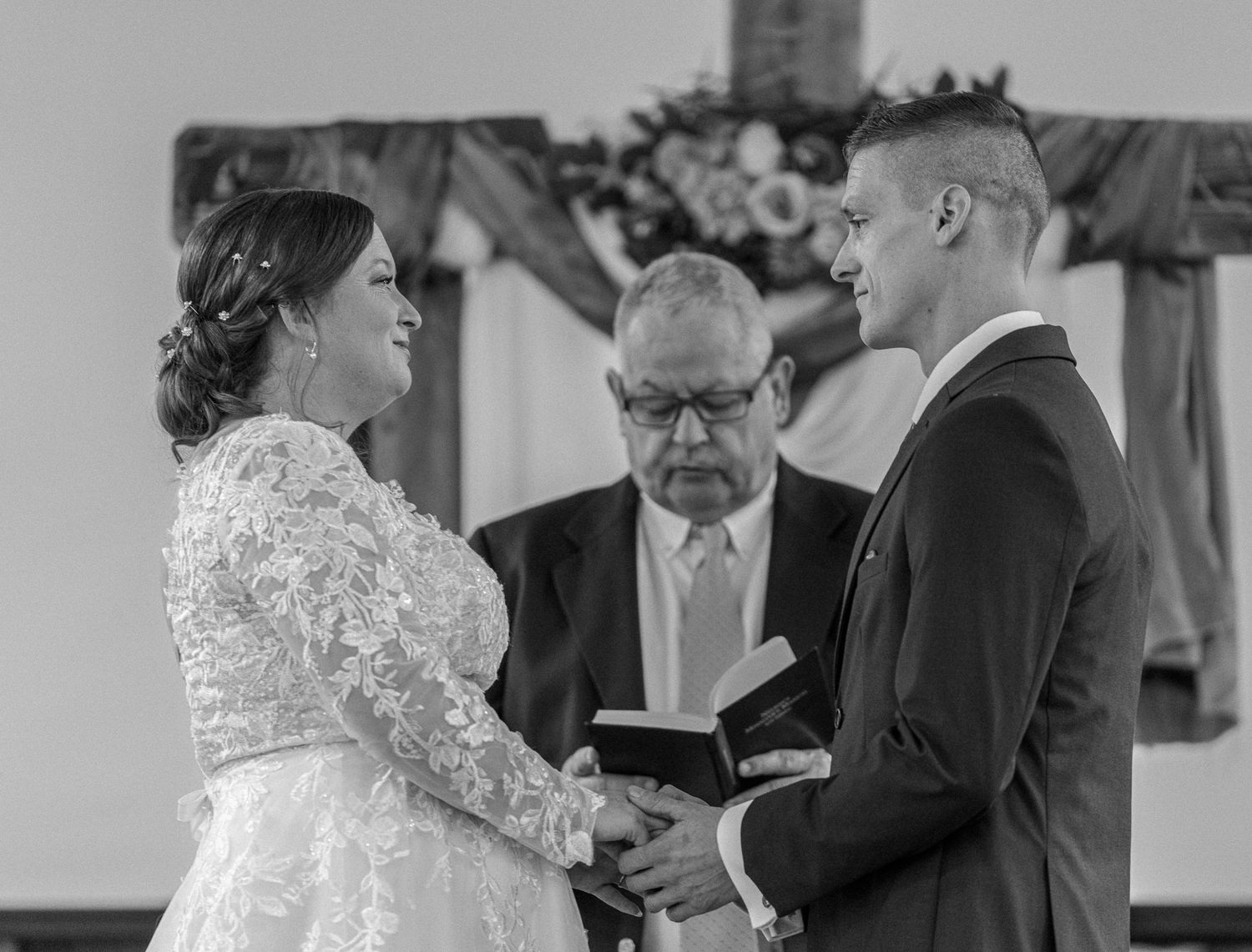 Black and white wedding ceremony photo of bride and groom exchanging vows before officiant in church.