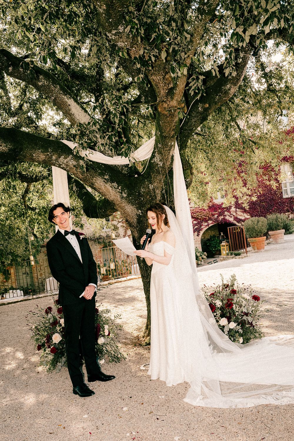 Bride and groom stand under a large oak tree with white draping during an outdoor wedding ceremony.