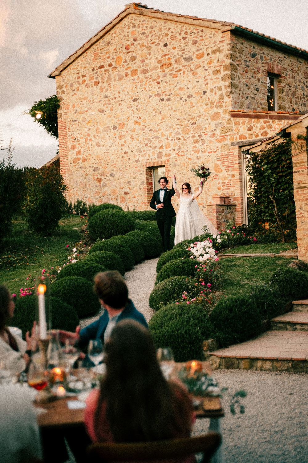 Bride and groom celebrate outside a rustic Tuscan stone villa at dusk, with guests dining al fresco by candlelight.