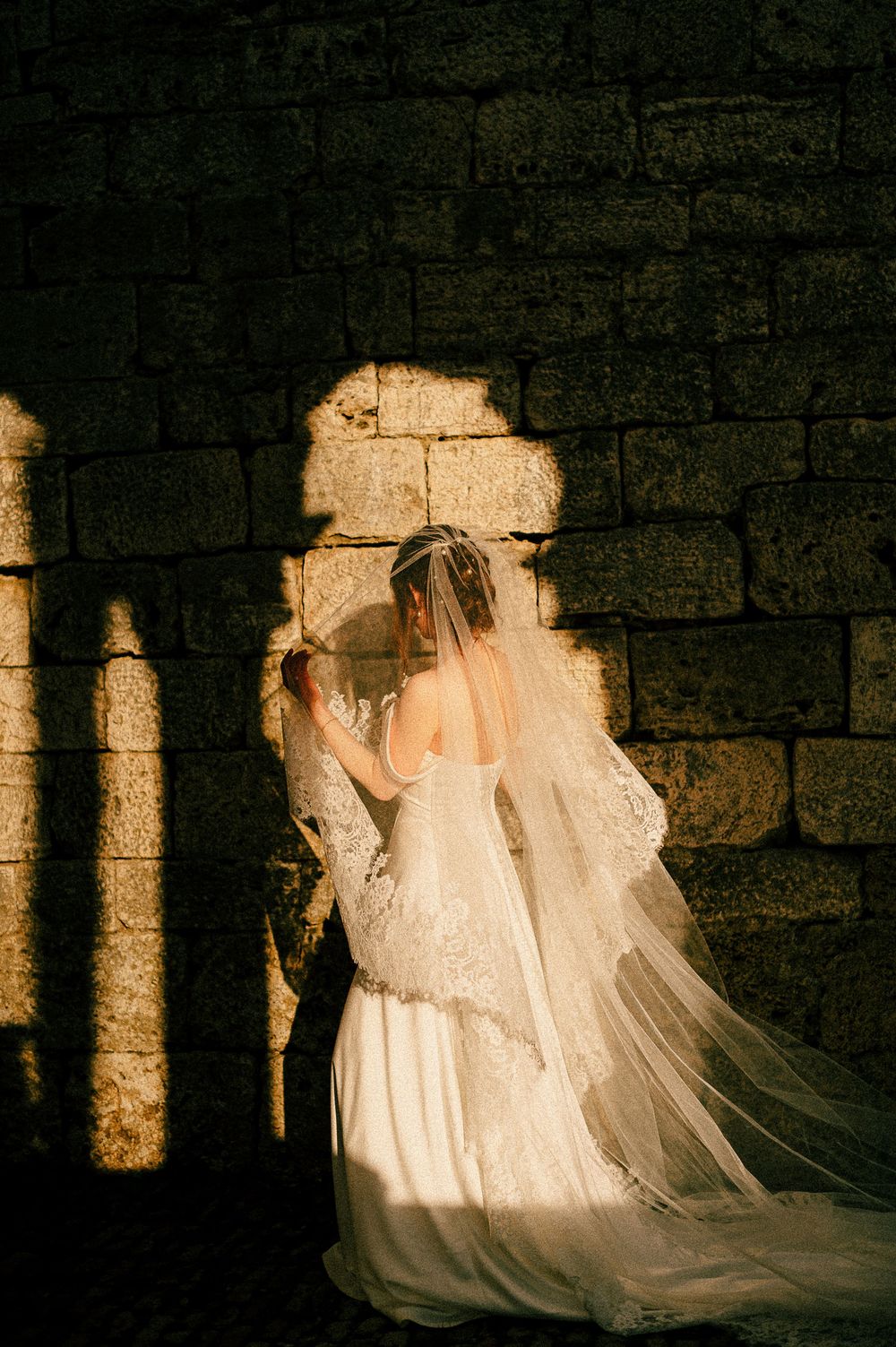Bride in lace wedding dress and flowing veil stands against stone wall in dramatic golden light.