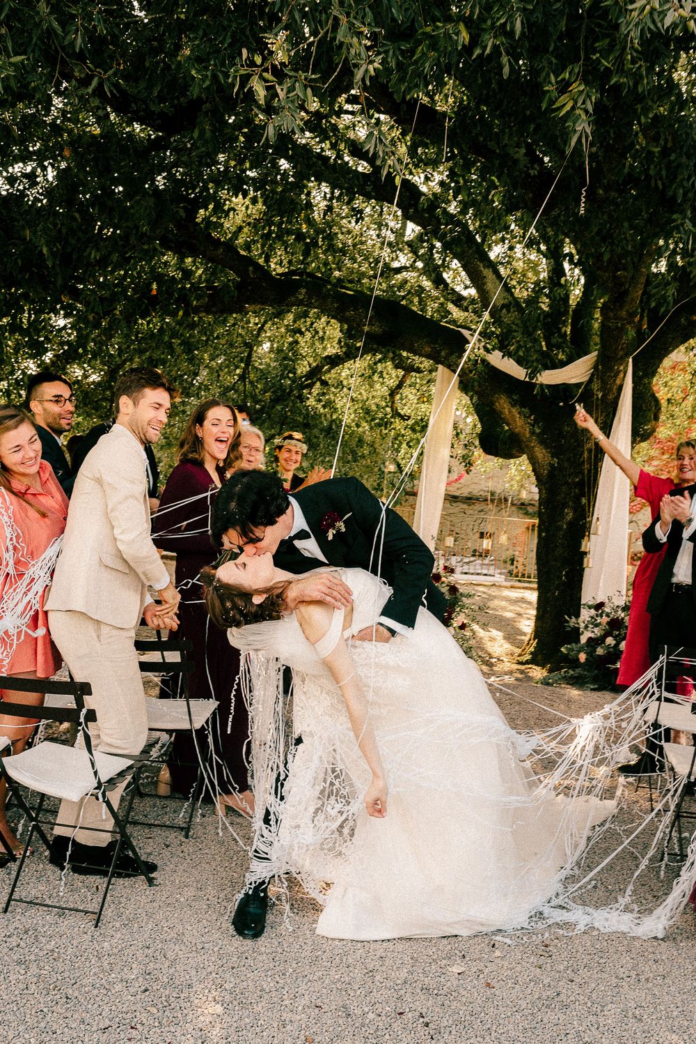 A bride and groom share a romantic dip kiss at their outdoor wedding ceremony surrounded by cheering guests under a large tree.