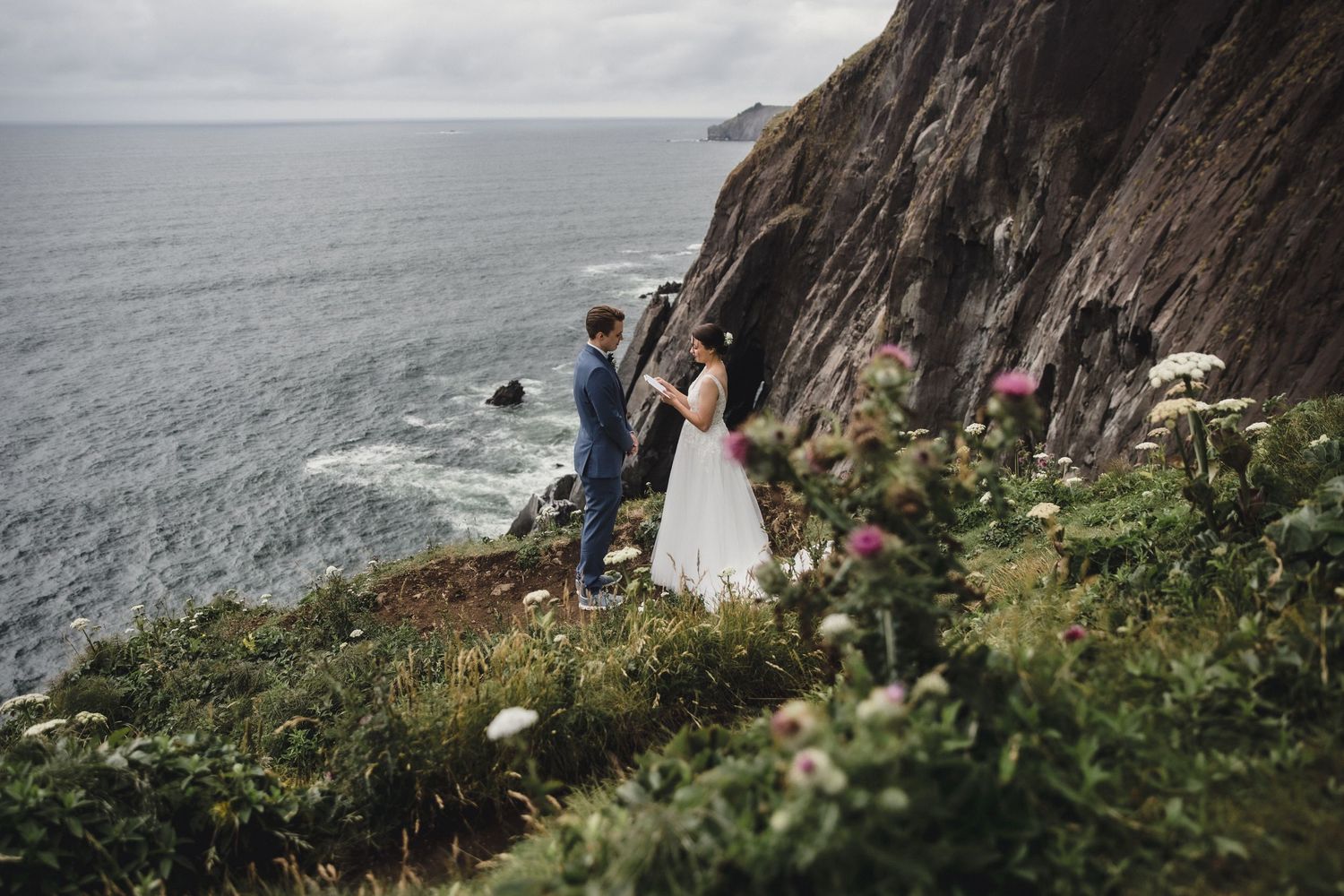 Bride and groom hold hands near a dramatic rocky sea cave on a pebble beach during their elopement.