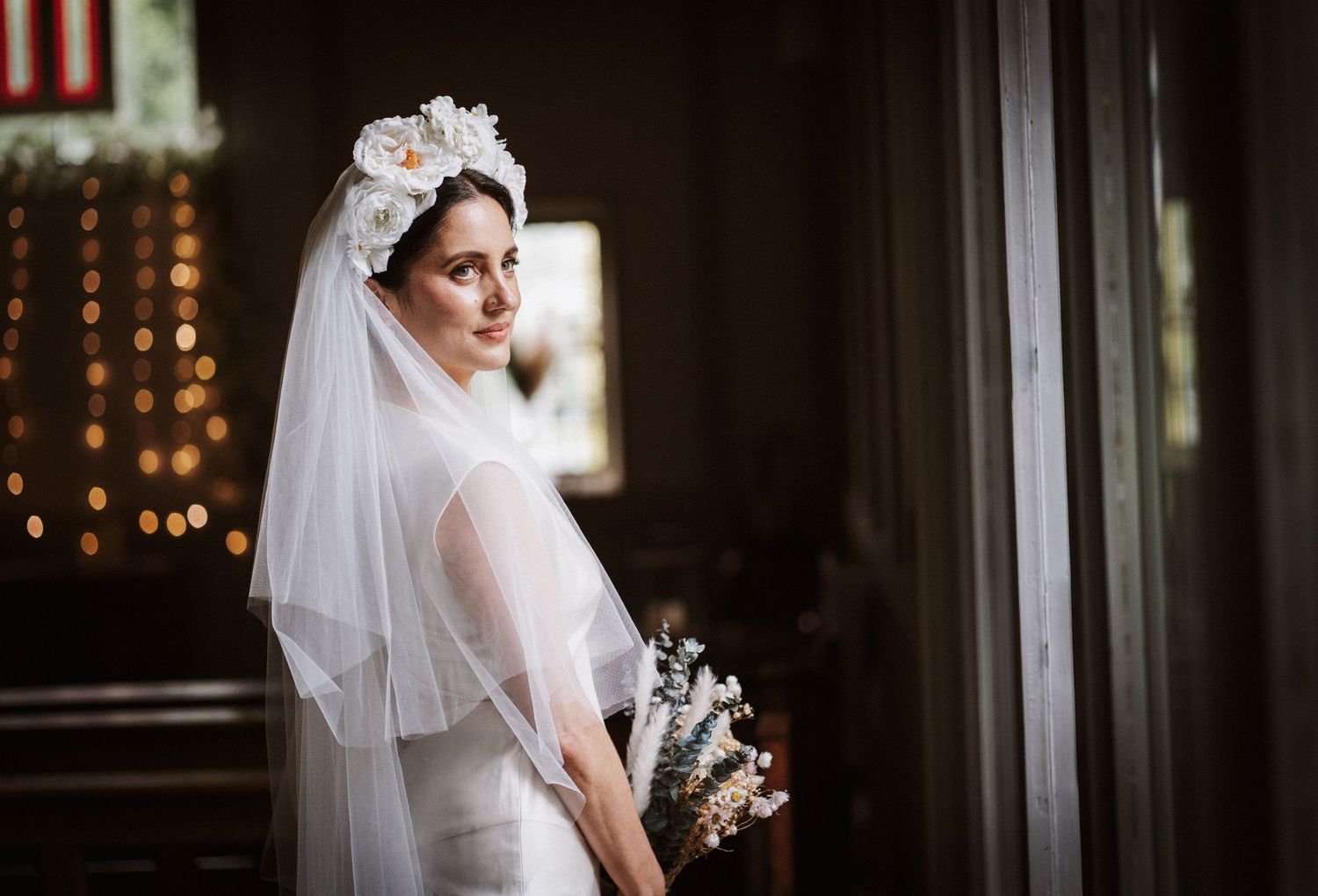 Elegant bride in white gown and floral veil holding bouquet, standing in soft natural light inside a church.