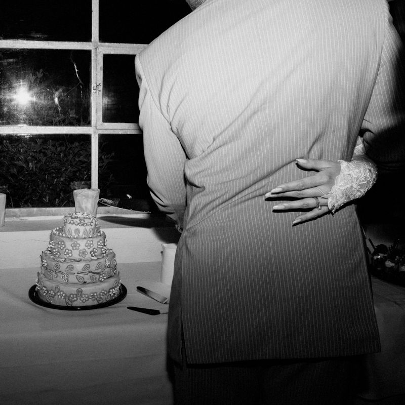 Black and white photo of a couple embracing near a tiered wedding cake at a reception.