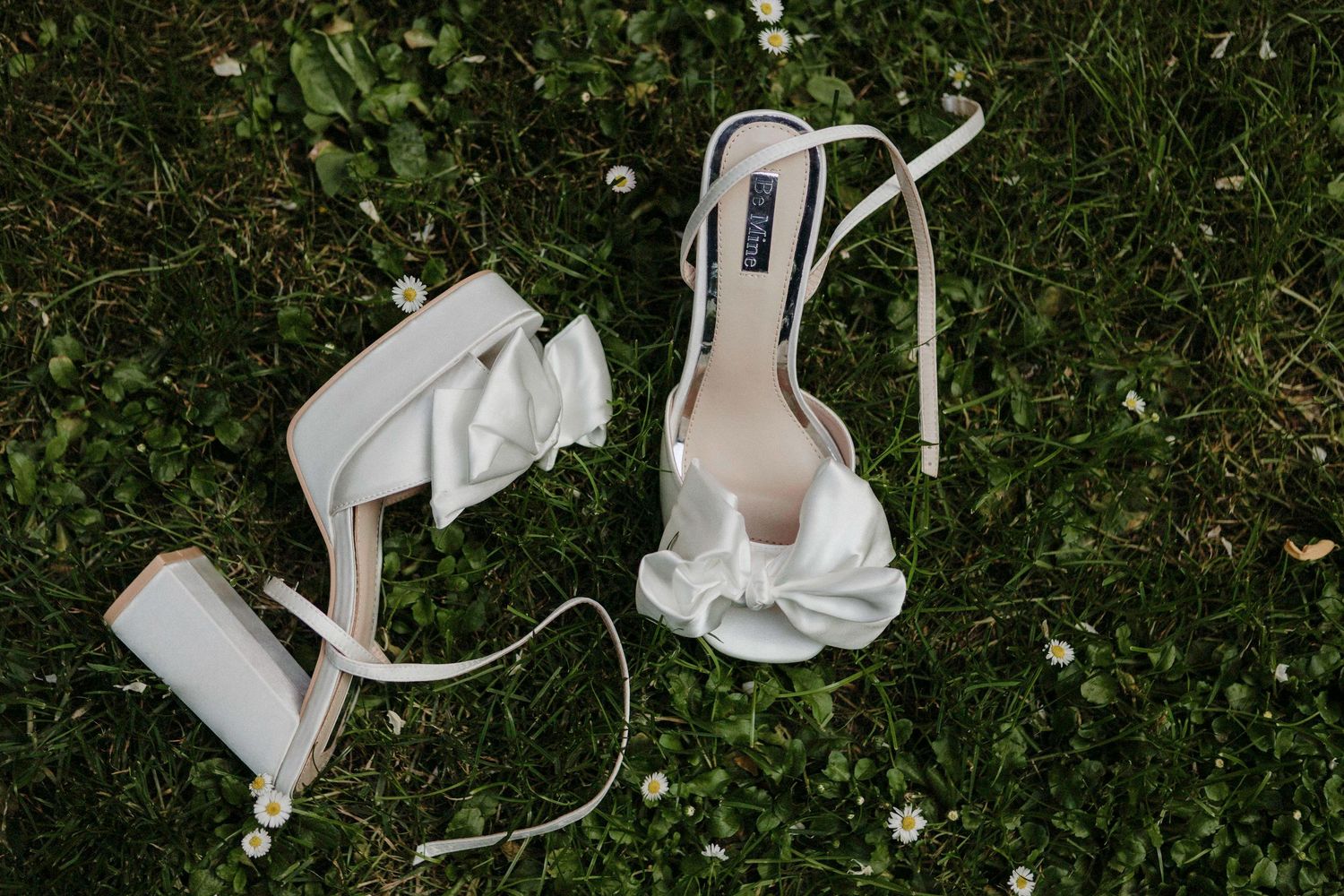 White satin bridal heels with bow detail and ankle ties resting on green grass with small white flowers.
