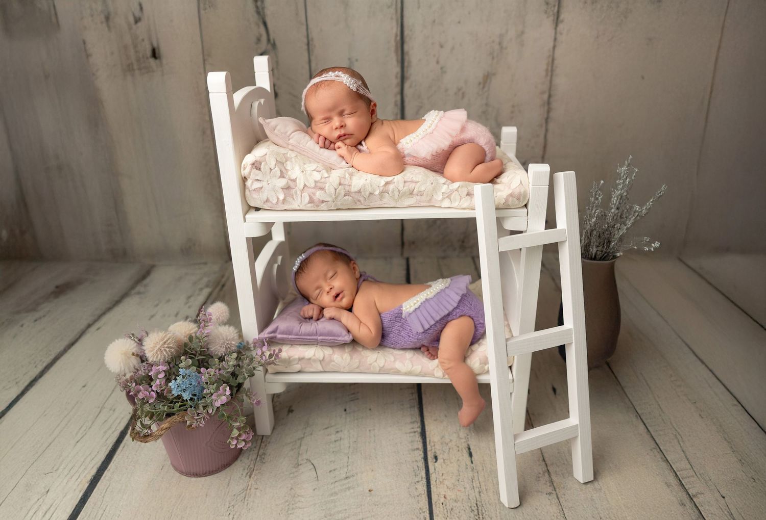 A sleeping newborn baby in a cream-colored bowl surrounded by pink flowers and greenery in a studio setting.