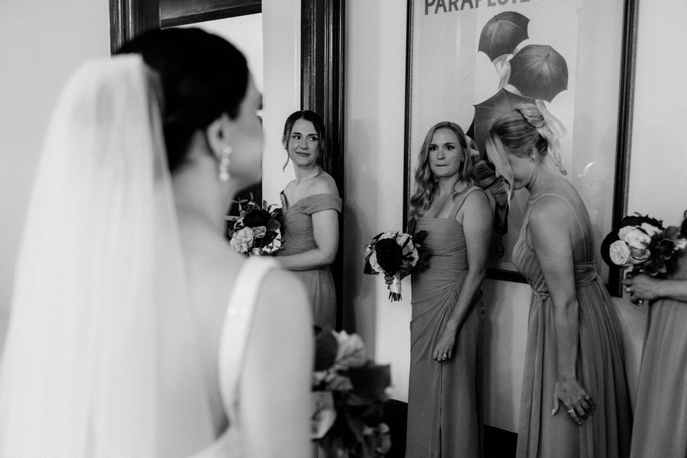 Photojournalistic wedding photo of bridesmaids in gowns lined up before ceremony with vintage poster backdrop.