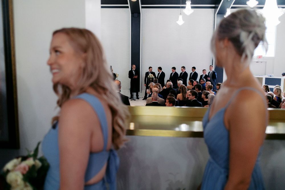 Two bridesmaids in blue dresses stand at a wedding ceremony with groomsmen and seated guests visible in the background.