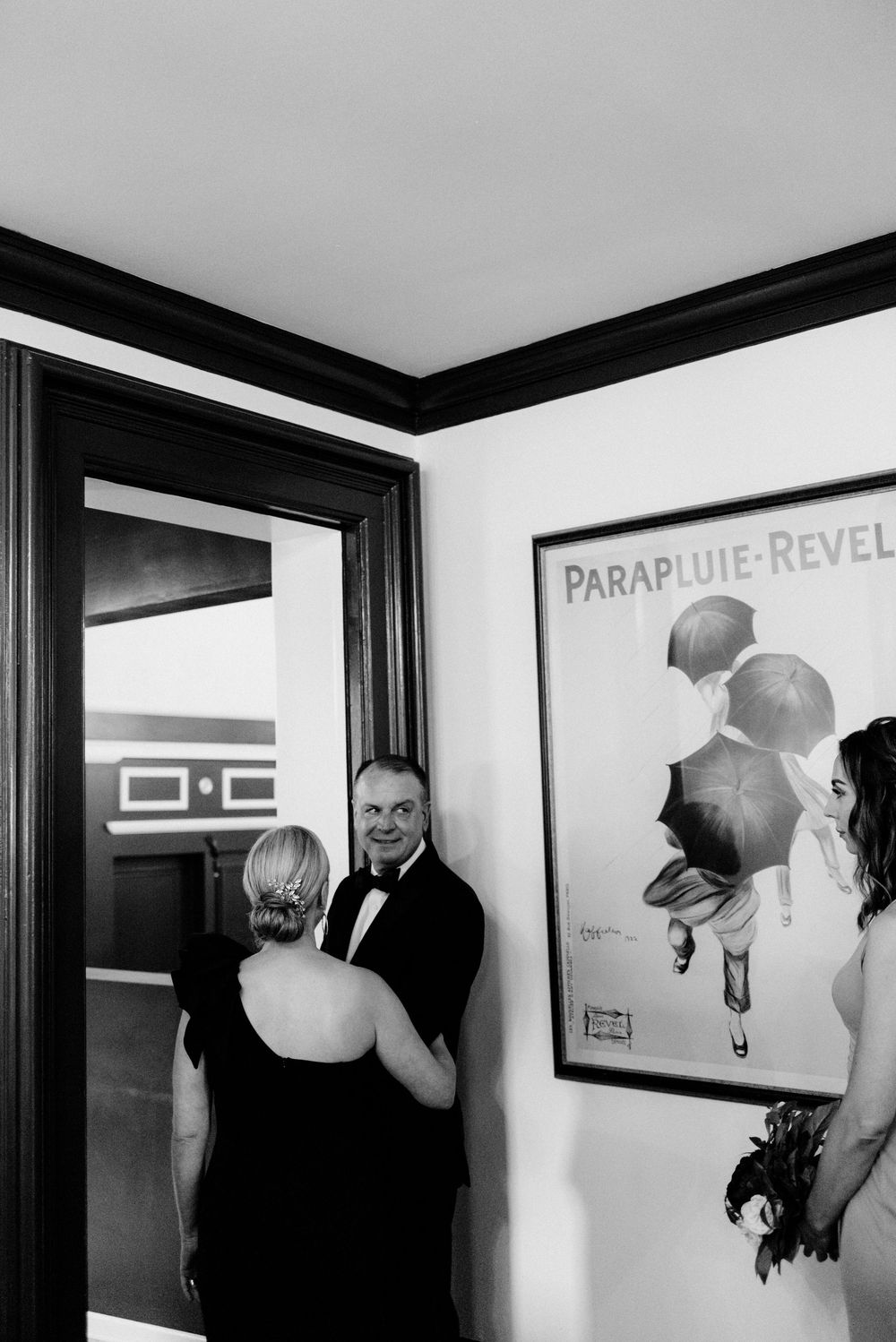 Black and white wedding photo of father and mother of the bride near a Parapluie Revel poster in an elegant interior.