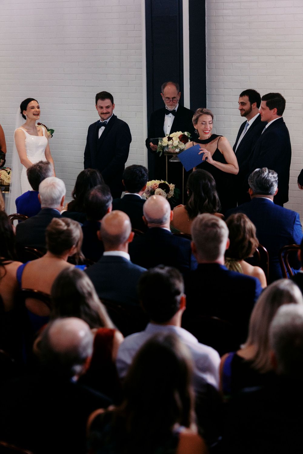 Wedding ceremony with bride, groom, officiant, and guests gathered in an elegant venue with white brick walls.