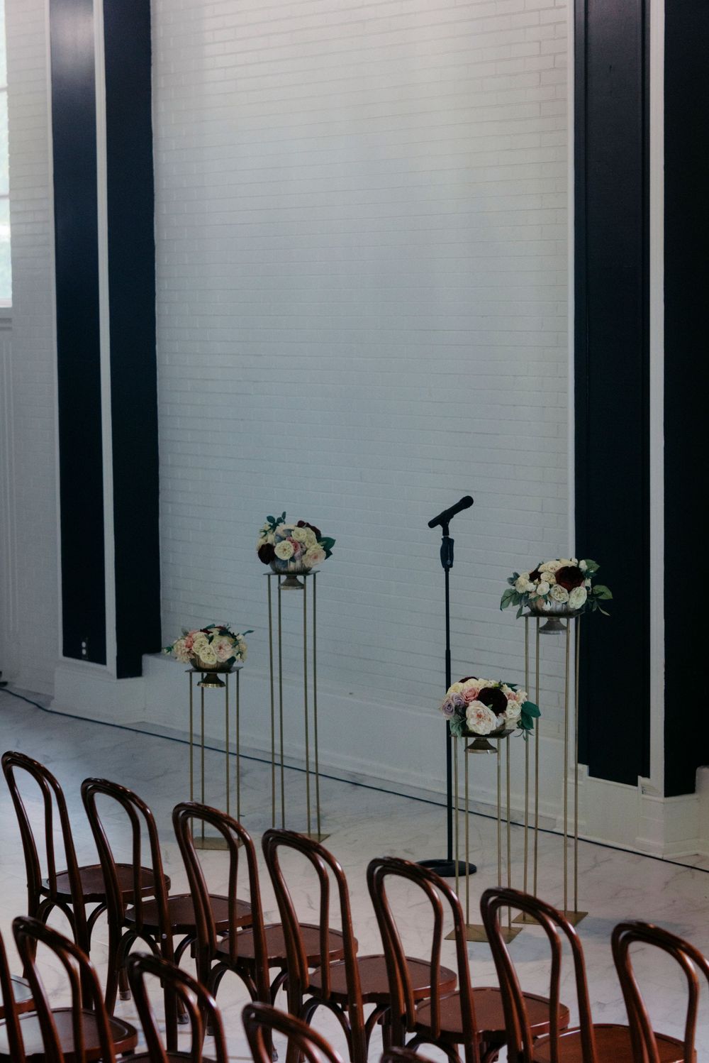 Elegant wedding ceremony setup with floral arrangements on tall stands, dark wood chairs, and a microphone against a white brick wall.