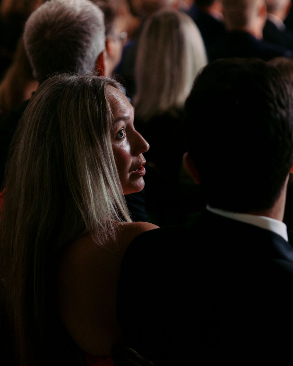 A woman with long blonde hair in profile view, seated in a dimly lit audience at a wedding ceremony.