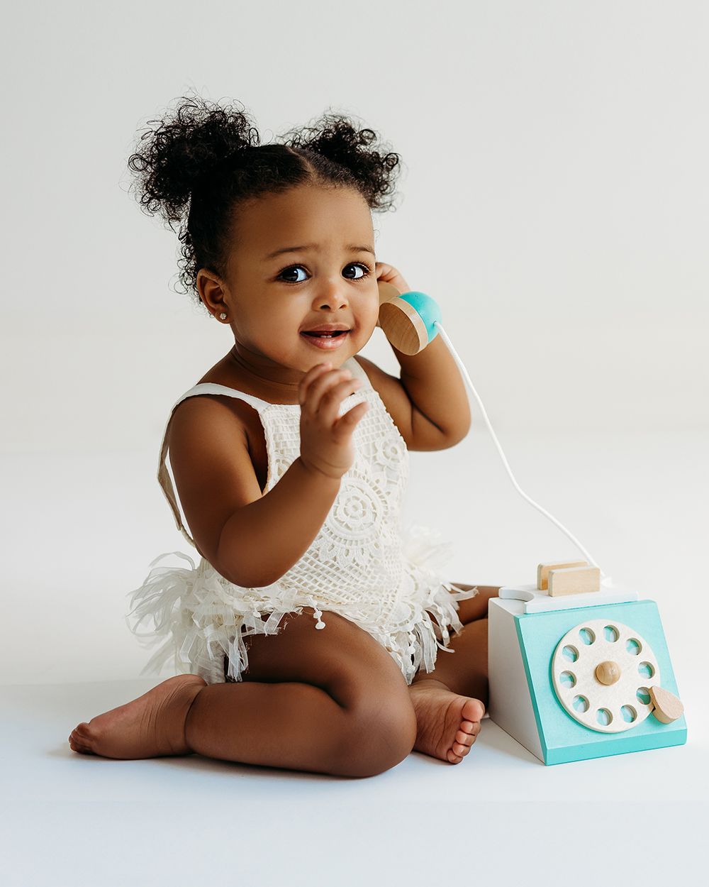 Adorable baby girl in white lace romper holds toy phone receiver, sitting beside a turquoise rotary toy telephone.