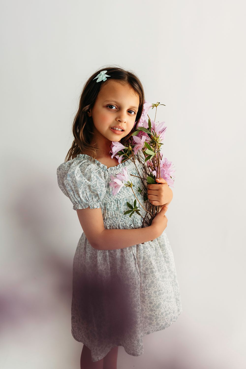 Young girl in floral dress holding pink wildflowers, posing against white background with butterfly hair clip.