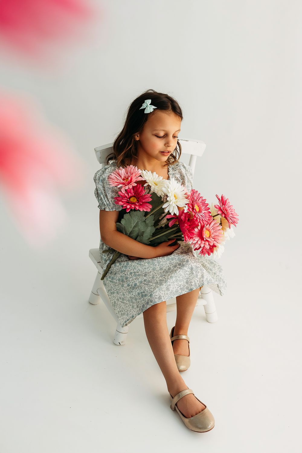 Young girl in floral dress sitting on white chair holding pink and white gerbera daisy bouquet in studio.
