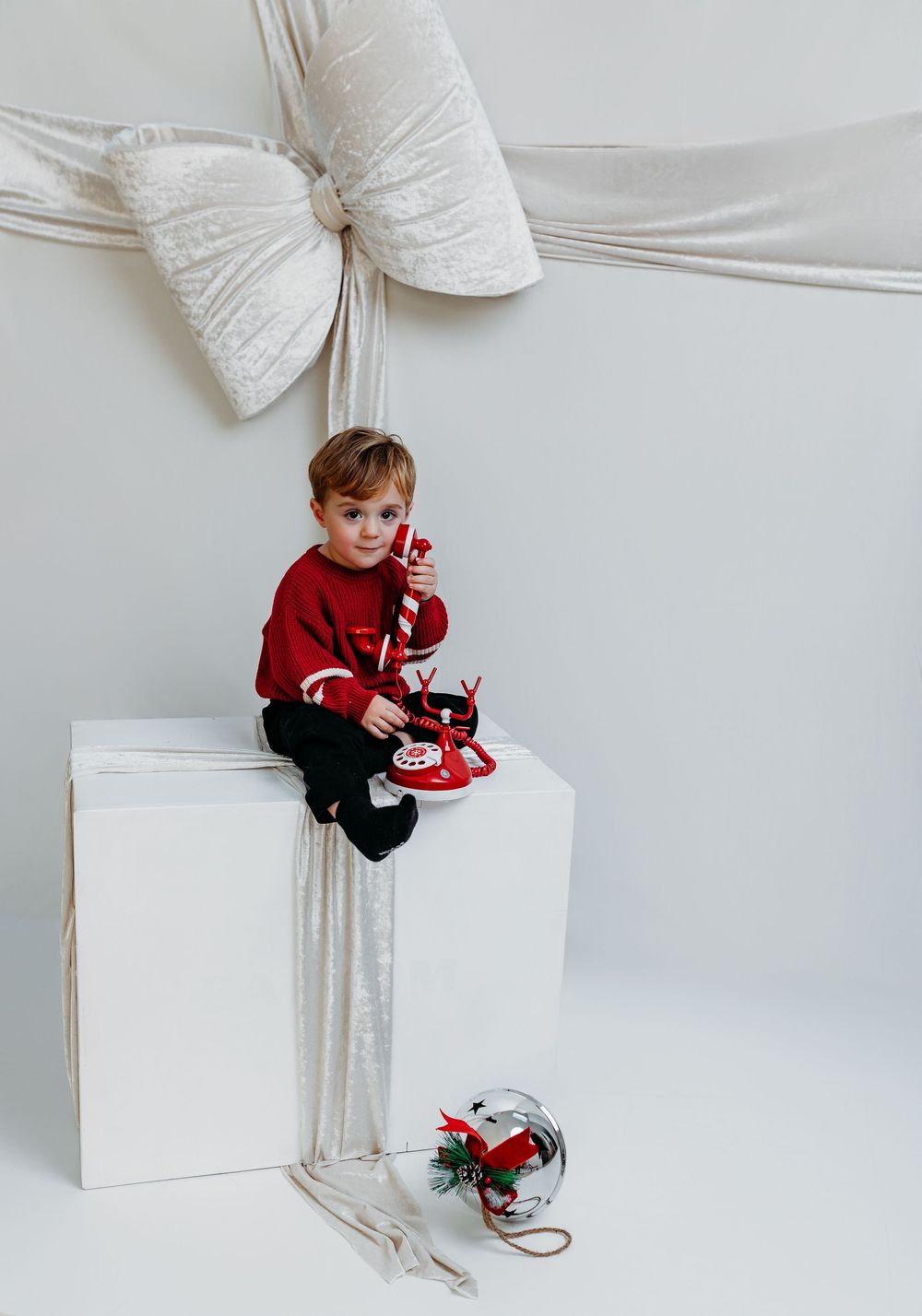 Young boy in red sweater sitting on white box with Christmas decorations and large bow backdrop.