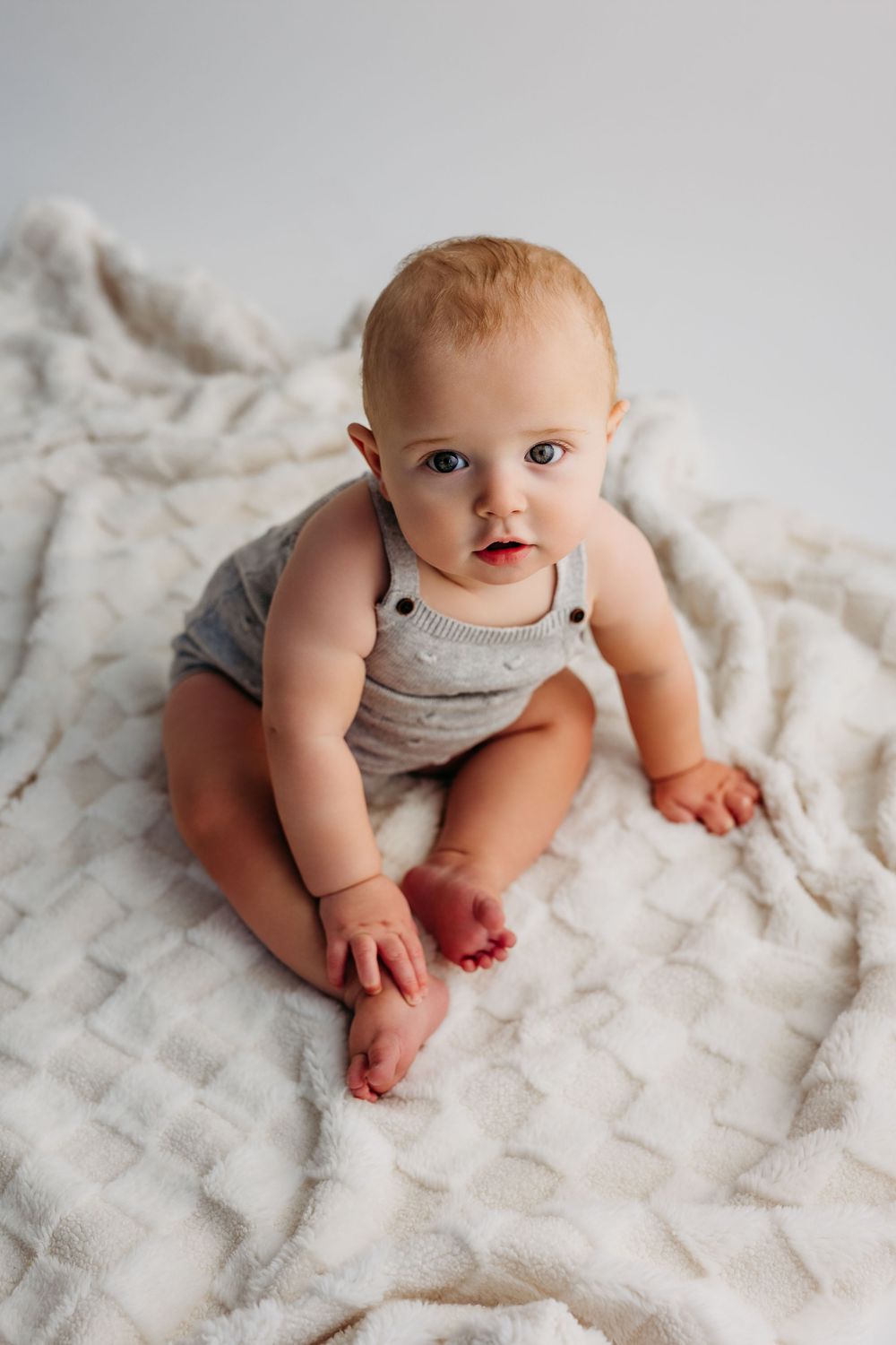 Adorable baby in gray overalls sitting on a soft cream textured blanket, looking up with bright blue eyes.
