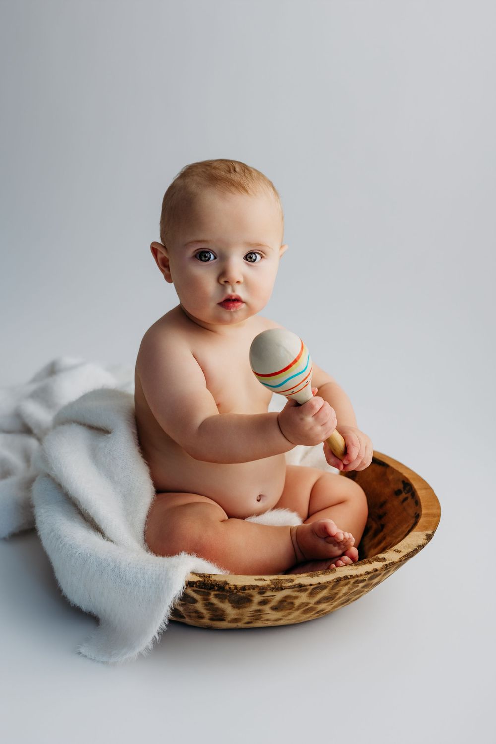 Cute baby sitting in a wooden bowl holding a colorful wooden maraca toy, wrapped in a white blanket on white background.