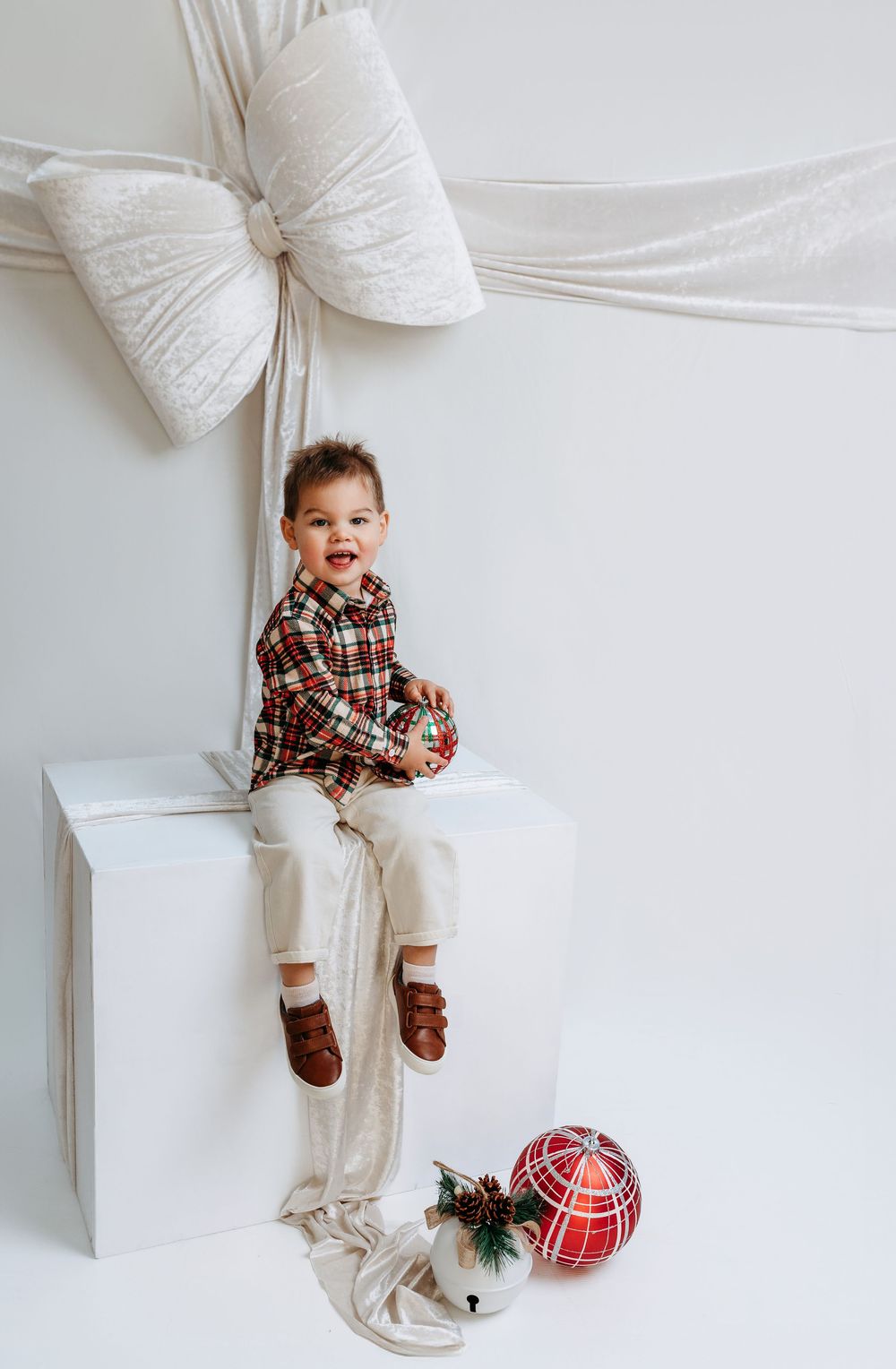 A smiling toddler in plaid shirt sits on white gift box with large bow, surrounded by Christmas ornaments.